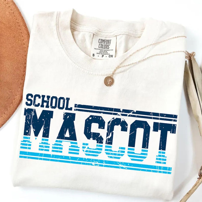 May include: Cream-colored t-shirt with the words "SCHOOL MASCOT" in distressed navy blue and light blue lettering. The shirt is folded and has a Comfort Colors tag.