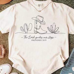 May include: Cream-colored t-shirt featuring a black line drawing of a cowboy boot, hat, and cacti. The text "The Lord guides our steps - PROVERBS 16:9" is printed below the graphic. A casual, country-themed design.