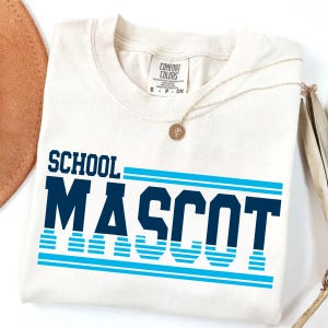 May include: Cream-colored t-shirt with the words "SCHOOL MASCOT" in navy blue block letters and light blue stripes. A gold necklace with a pendant rests on the shirt.