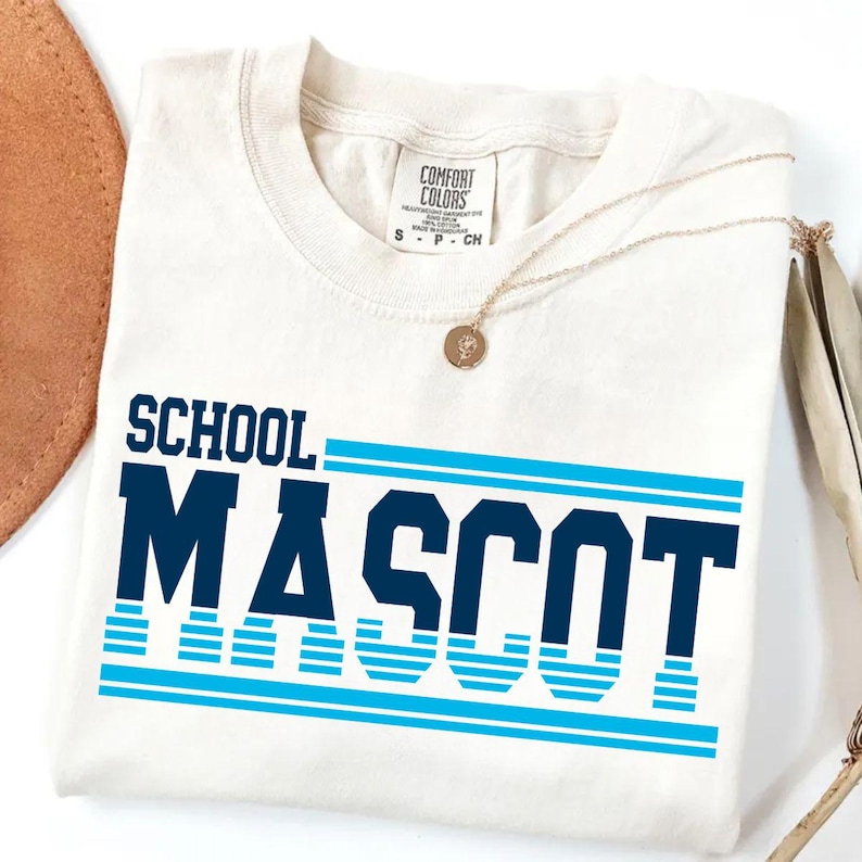 May include: Cream-colored t-shirt with the words "SCHOOL MASCOT" in navy blue block letters and light blue stripes. A gold necklace with a pendant rests on the shirt.