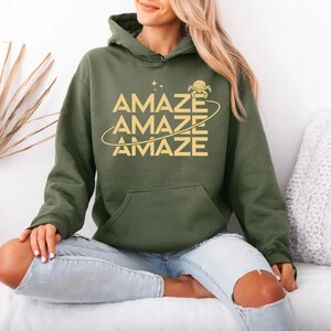 Op de afbeelding: Olijfgroene hoodie met het woord "AMAZE" in goud, drie keer gestapeld. Het ontwerp bevat een planeetgrafiek met een ring en sterren. De hoodie heeft een voorzak en een capuchon.