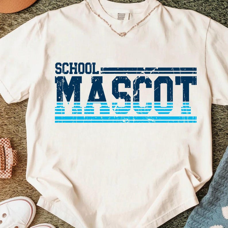 May include: A white t-shirt with the words "SCHOOL MASCOT" printed in blue. The text is in a bold, distressed font, with a horizontal blue stripe design. The shirt has a crew neck.