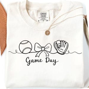 Op de afbeelding: Crèmekleurig T-shirt met een zwarte lijntekening van een honkbal, een strik en een honkbalhandschoen. De woorden "Game Day" staan onder de tekening in een scriptlettertype. Een gouden ketting met een hanger is over het shirt gedrapeerd.