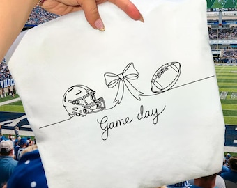May include: White pillow with a black line drawing of a football helmet, bow, and football. The words "Game day" are written in a script font below the drawings. The pillow is held up in front of a football stadium.