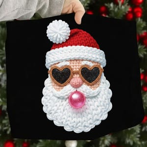 May include: A black fabric with a crocheted Santa Claus face. Santa wears heart-shaped sunglasses with black glitter lenses, a red hat with a white pom-pom, and a white beard. A pink bubblegum-like orb is below the nose.