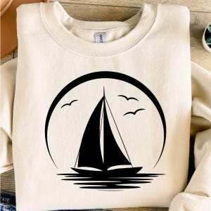 May include: A cream-coloured sweatshirt with a black sailboat design. The design features a sailboat on the water, set against a circular frame, with three birds flying above. The sail is triangular.