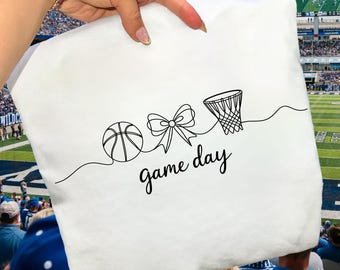May include: White square pillow with a black line drawing of a basketball, bow, and basketball hoop. The words "game day" are written in a script font below the drawings. The pillow is held up by a person.