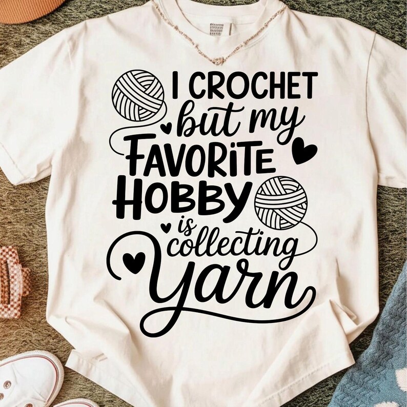 May include: Cream-colored t-shirt with the text "I Crochet but my FAVORITE HOBBY is collecting yarn" in black script and block letters. The design includes yarn ball illustrations and heart accents.