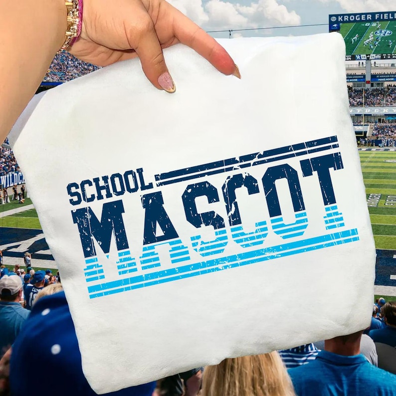 May include: White pillow with the words "SCHOOL MASCOT" in distressed navy blue and light blue lettering. The pillow is held up in front of a football stadium.