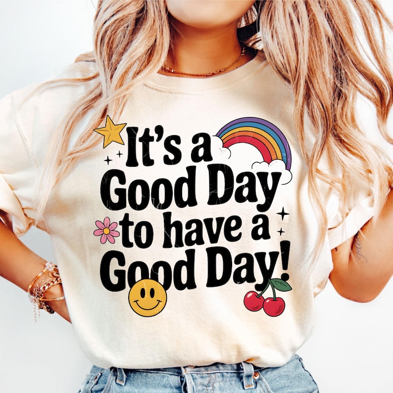 May include: A white t-shirt with the text 'It's a Good Day to have a Good Day!' in black. The text is surrounded by colorful graphics including a yellow star, a pink flower, a rainbow, a cloud, a smiley face, and two red cherries.
