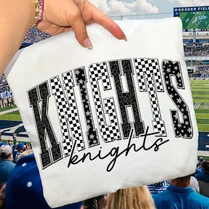 May include: White t-shirt with the word "KNIGHTS" in large, bold letters, featuring a mix of checkered, leopard print, and star patterns. The word "knights" is written in a cursive font below the main text. The shirt is held up in front of a stadium.