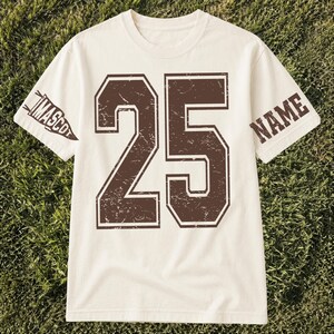 Puede incluir: Una camiseta de manga corta color crema con un número 25 grande de color marrón desgastado impreso en el centro. La manga izquierda muestra un gráfico de banderín marrón con la palabra MASCOT, y la manga derecha tiene la palabra NAME en marrón.