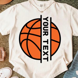 Peut inclure: T-shirt blanc cass&eacute; avec un motif de ballon de basket-ball. Le ballon est orange avec des lignes noires, et un rectangle noir vertical sur le c&ocirc;t&eacute; droit affiche le texte "YOUR TEXT" en noir. Le t-shirt a un col rond.
