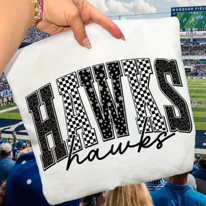 May include: White pillow with the word "HAWKS" in large, bold letters. The letters have a mix of patterns, including leopard print, checkered, and star designs. The word "hawks" is written in a cursive font below the main word.