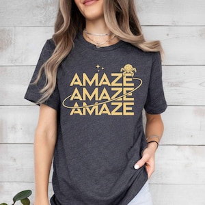 Op de afbeelding: Donkergrijs t-shirt met het woord "AMAZE" driemaal in goud. Het ontwerp bevat een gestileerde planeet en sterren. Het shirt heeft een ronde hals en korte mouwen.