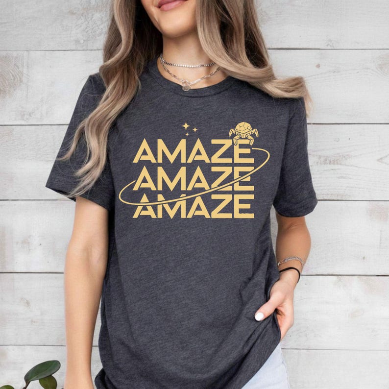 Op de afbeelding: Donkergrijs t-shirt met het woord "AMAZE" driemaal in goud. Het ontwerp bevat een gestileerde planeet en sterren. Het shirt heeft een ronde hals en korte mouwen.