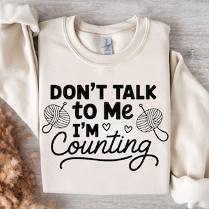 May include: Cream-colored sweatshirt with the text "DON'T TALK to Me I'M Counting" in black. The design includes yarn balls and hearts, suggesting a knitting or crafting theme. A cozy and comfortable garment.
