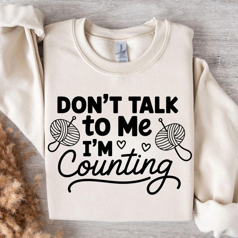 May include: Cream-colored sweatshirt with the text "DON'T TALK to Me I'M Counting" in black. The design includes yarn balls and hearts, suggesting a knitting or crafting theme. A cozy and comfortable garment.