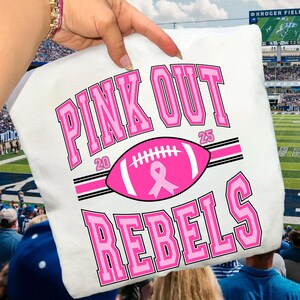 Może przedstawiać: Biała koszulka z napisem "PINK OUT REBELS" w różowych literach. Różowa piłka nożna amerykańska z różową wstążką jest umieszczona na środku koszulki. Numery "20" i "25" znajdują się po obu stronach piłki.