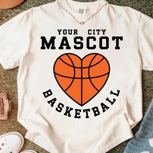 Peut inclure: T-shirt blanc avec l'inscription noire "YOUR CITY MASCOT". En dessous, un motif de ballon de basket-ball en forme de c&oelig;ur orange et noir, avec le mot "BASKETBALL" en arc de cercle en bas.