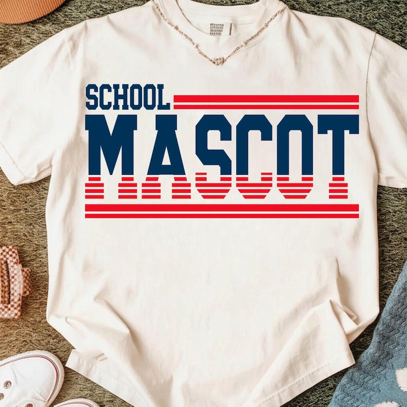 May include: A white t-shirt with the words "SCHOOL MASCOT" printed in navy blue and red. The text is arranged in a bold, blocky font, with the word "MASCOT" prominently displayed. The shirt is made of a soft material.