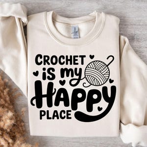 May include: Cream-colored sweatshirt with the text "CROCHET is my HAPPY PLACE" in black. The design includes a yarn ball and hearts. A cozy, casual top for crafters.