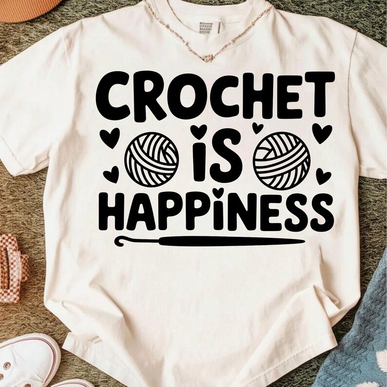 May include: Cream-colored t-shirt with the text "CROCHET IS HAPPINESS" in bold black letters. The design includes yarn balls, hearts, and a crochet hook. A casual, comfortable top for crafters.