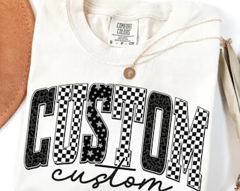 May include: A cream-colored t-shirt featuring the word CUSTOM in large block letters with alternating black and white checkerboard, leopard print, and star patterns, with the word custom written in black cursive script underneath the block text.