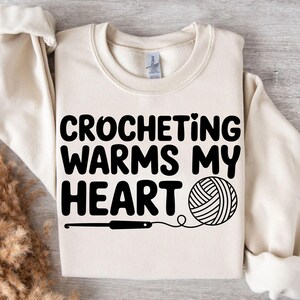 May include: Cream-colored sweatshirt with the text "CROCHETING WARMS MY HEART" in bold black letters. A black crochet hook and a yarn ball graphic are below the text, representing the craft of crocheting.