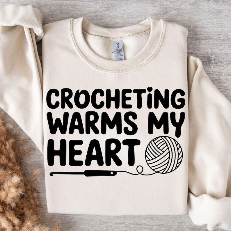 May include: Cream-colored sweatshirt with the text "CROCHETING WARMS MY HEART" in bold black letters. A black crochet hook and a yarn ball graphic are below the text, representing the craft of crocheting.