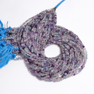 May include: A collection of faceted gemstone beads in shades of purple, lavender, and clear, strung together. The beads are oval-shaped and appear to be made of fluorite. A blue tassel is attached to the top of the beads.
