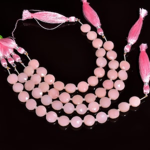 May include: Strands of light pink gemstone beads with faceted surfaces. Each strand is strung with small white beads and finished with a pink tassel. The beads are arranged on a black background.