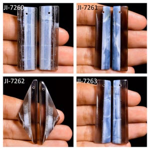 Puede incluir: Cuatro juegos de colgantes de piedra pulida etiquetados como JI-7260, JI-7261, JI-7262 y JI-7263. Cada colgante presenta una banda vertical de piedra azul translúcida flanqueada por bordes de color marrón ahumado oscuro. Las piezas son rectangulares o en forma de diamante con un pequeño orificio perforado en la parte superior para colgar.