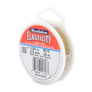 Op de afbeelding: Een spoel Beadalon Elasticity elastisch koord in een witte plastic houder. Het etiket is rood en wit, met tekst die aangeeft dat het koord 0,5 mm in diameter is en 25 m lang. Het koord is satijn goudkleurig.
