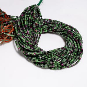 May include: Strands of green and purple gemstone beads. The beads are small, square-cut, and strung together. The color palette includes shades of green, purple, and a hint of silver. The beads are likely used for jewelry making.