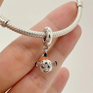 May include: A silver charm bracelet with a dangling Nemo charm. The charm features a smiling orange and white clownfish with black accents. The bracelet is made of silver and has a textured, snake-like design.