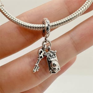 May include: A silver charm featuring a key and a small, detailed box pendant. The charm is attached to a silver bracelet. The key has a decorative handle, and the box pendant has a rounded top and a small, round detail.