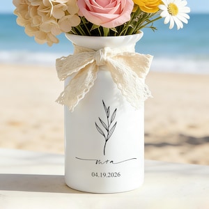 Pode incluir: Vaso de cerâmica branca com um arranjo floral de rosas cor-de-rosa, margaridas amarelas e brancas e flores creme. O vaso tem um design botânico preto e a data 04.19.2026. Uma fita de renda creme adorna o vaso.