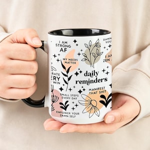 May include: White ceramic mug with a black interior and handle, featuring the text "daily reminders" and other motivational phrases in black. The mug is decorated with floral and star graphics in black, peach, and beige.