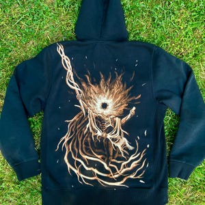 Puede incluir: Sudadera con capucha negra con una ilustración detallada en la espalda. La obra de arte representa una figura esquelética con un bastón, rodeada de un diseño ardiente y radiante en tonos marrones y beige. La sudadera con capucha está sobre hierba verde.