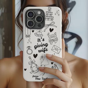May include: White phone case featuring black line art illustrations of wedding symbols, such as champagne flutes, a ring, and a car. The phrase "It's giving wife" is written in a script font. The words "future Mrs. Taylor" are also included.