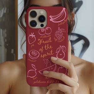 May include: A maroon phone case featuring a design of various fruits and the words "Fruit of the Spirit" in a handwritten style. The case includes illustrations of a banana, grapes, a pear, a lemon, and other fruits. The text "galatians 5:22-23" is also visible.
