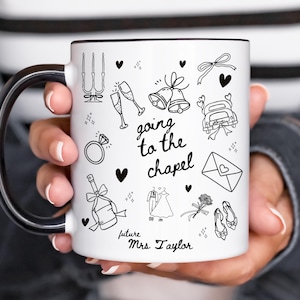 May include: White ceramic mug with a black handle and rim, decorated with black line art illustrations of wedding-related icons, including champagne glasses, bells, and a wedding dress. The text "going to the chapel" and "future Mrs. Taylor" are also featured.