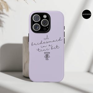 May include: A lavender phone case with a black camera module. The case features the text "A bridesmaid in a 'tini bit" in a handwritten font, along with a small illustration. A black circle with the word "LAVENDER" is in the upper right corner.