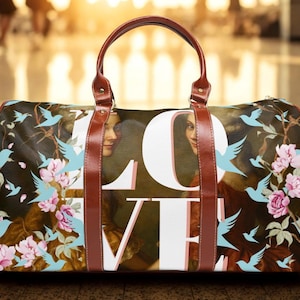 Può includere: Una borsa da viaggio con manico e finiture in pelle marrone. La borsa presenta un motivo floreale e di uccelli con la parola "LOVE" a grandi lettere bianche. Lo sfondo presenta un dipinto di una donna.