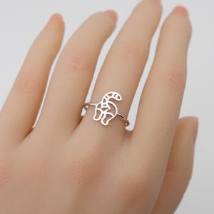 May include: A silver ring featuring a cut-out design of a cat's rear end. The ring is worn on a finger, showcasing the delicate details of the feline-inspired jewelry. The ring is a simple, yet playful accessory.