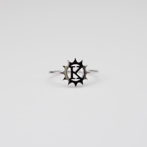 Puede incluir: Un anillo plateado con un diseño de sol y la letra "K" en el centro. El anillo se muestra sobre un fondo blanco liso, resaltando los intrincados detalles de la joya.