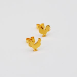 Puede incluir: Un par de pendientes de botón de gallo de color dorado. Los gallos están mirando hacia la derecha y tienen un diseño detallado.