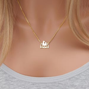 May include: A gold chain necklace with a pendant featuring two birds perched on an open book. The pendant is a delicate, cutout design.