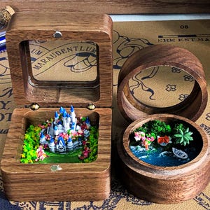 May include: Two wooden boxes with glass lids. One box contains a miniature castle with blue and white details, surrounded by green and pink flowers. The other box contains a miniature scene with a blue lake, green trees, and a small house.